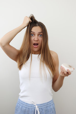 Shocked young woman with hair loss problem holds on to the hair and hairbrush with a bunch of fallen hair after combing. Postpartum period, menstrual or endocrine disorder, hormonal imbalance, stressの写真素材