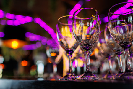 Empty clear wineglasses stand in rows on bar counter at nightclub, blurred purple neon and yellow lights on background. Clean cocktail glasses ready to celebrate party.の写真素材