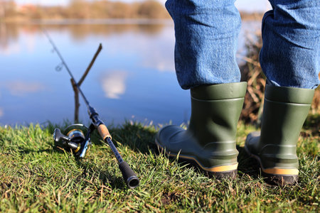 Cropped photo of fisherman's legs in rubber boots with rod, spinning reel on river bank....