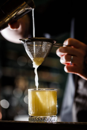 man bartender carefully filters bright alcoholic cocktail from steel shaker cup into glass through...