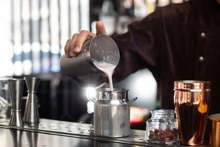 Barman gently pours finished cocktail from glass shaker into metalic glass. Body of bartender in brown apron on backgroundの写真素材