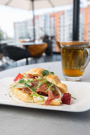 Croissant sandwich with prosciutto, berries, dijon mustard, cheese and cup of tea on white plate and bright background. Continental style breakfastの写真素材