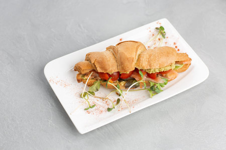 Croissant sandwich with avocado, tomatoes on white plate and bright background. Continental style...