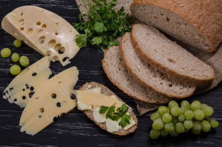 Rural breakfast. Cheese, bread, parsley and grape on the black wooden table. Cheese and bread are...