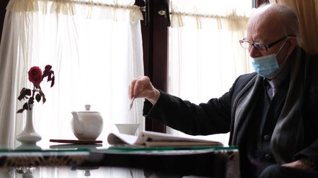 An old retiree in a protective mask stirs tea while sitting in a cafe