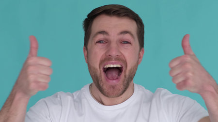 Young guy in a white T-shirt actively showing thumbs up on a turquoise background in the studio