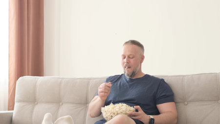 The man is relaxing at home on the couch, he is eating popcorn and watching TV. Happy relaxed guy