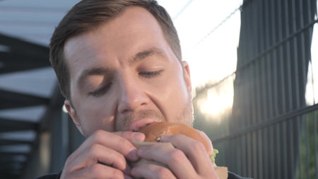 Businessman during lunch break eating hamburger or sandwich, close up, standing near business center