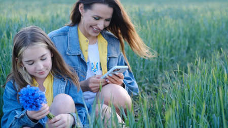 Mom and daughter use a smartphone sitting in the tall green grass in the middle of the field.の写真素材