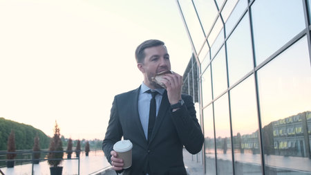 Young handsome businessman holding a cup of coffee, eating a burger on the street.の写真素材