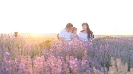 Beautiful young family dad and mom with little girl sitting on picnic at sunset in lavender field.の写真素材