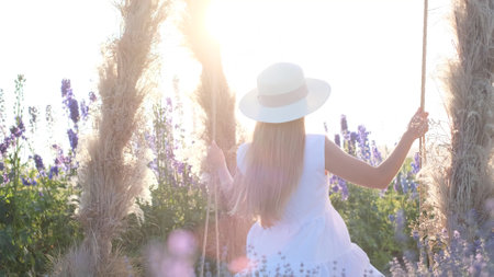 A young woman is riding on a swing at sunset, she is wearing a beautiful dress and a hat. Summerの写真素材