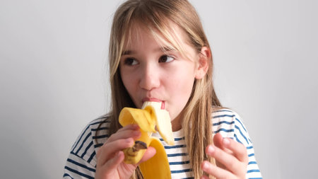 Happy little girl eating a yellow banana. A 7-year-old girl eats a banana on a white background.の写真素材