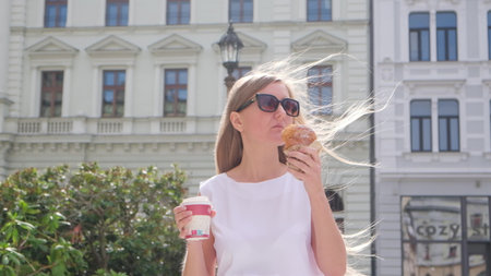 A young woman with a croissant and a cup of coffee on a city walk.