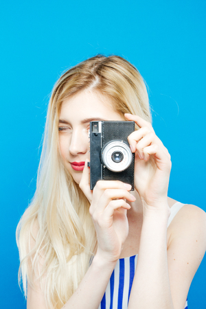 Cute Young Woman in Summer Striped Dress is Using Retro Camera on Blue Background in Studio.の写真素材