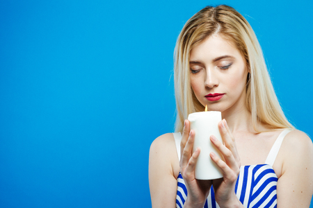 Portrait of Sad Serious Girl with Closed Eyes Holding White Candle in Her Hands. Beautiful Blonde with Bare Shoulders Wearing Striped Dress on Blue Background in Studio.の写真素材