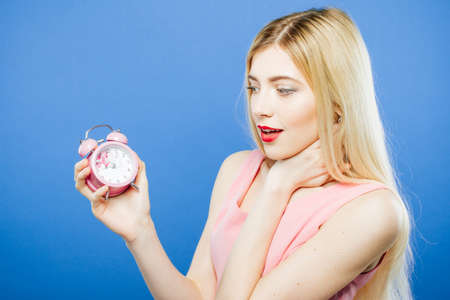Shocked Blonde Wearing Dress is Holding Pink Alarm Clock in Hand on Blue Background in Studio.の写真素材