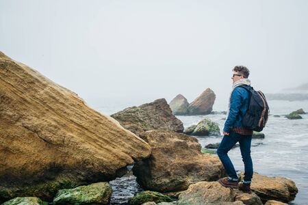 Traveler with a backpack stands on a rock against a beautiful sea with waves, a stylish hipster boy posing near a calm ocean during a wonderful journey around the world. Shoot from the backの写真素材