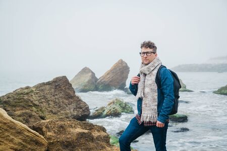 Man traveler with a backpack standing on a rock against a beautiful sea with waves, a stylish hipster boy posing near a calm ocean during a wonderful journey around the worldの写真素材