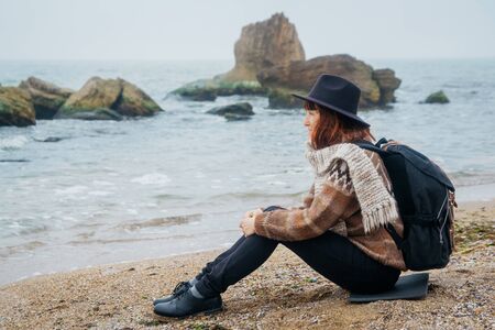 Young woman tourist in hat and with backpack sitting on beach, looking at sea, on coastline, on horizon. Tourism, vacation, lifestyle. Hipster girl resting on beachの写真素材