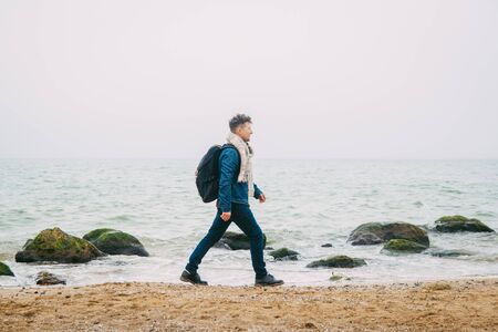 Traveler with a backpack standing near a rock against a beautiful sea with waves, a stylish hipster boy posing near a calm ocean during a wonderful journey around the world. Verticalの写真素材