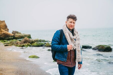 Traveler with a backpack standing near a beautiful sea with waves, a stylish hipster boy posing near a calm ocean during a wonderful journey around the world. A look at the cameraの写真素材