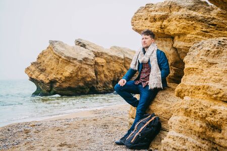 Young man traveler with a backpack standing on a rock against a beautiful sea with waves, a stylish hipster boy posing near a calm ocean during a wonderful journey around the worldの写真素材