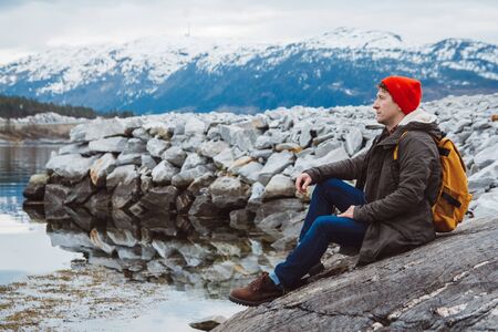 Traveler man with a yellow backpack wearing a red hat sitting on the shore on the background of mountain and lake. Space for your text message or promotional content. Travel lifestyle concept. Look to the side.の写真素材