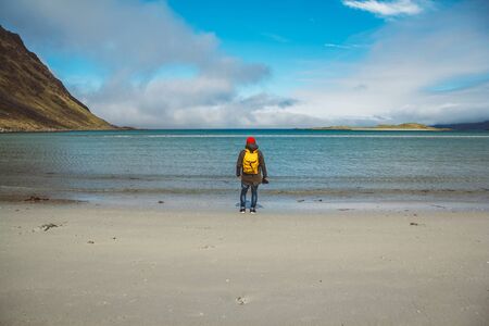 Traveler is a professional photographer taking over the landscape photo landscape. Wearing a yellow backpack in a red hat standing on a sandy beach on the background of the sea and mountains. Shoot from the back.の写真素材