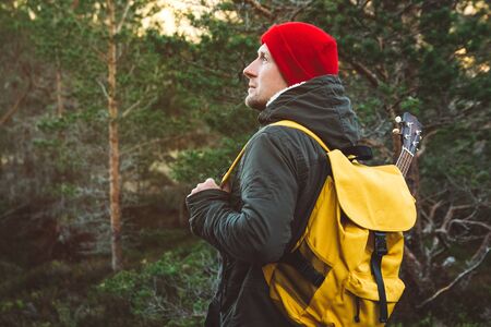 Traveler man is standing on a trail in the middle of a forest with a guitar. Wearing a yellow backpack in a red hat. Lifestyle Travel Concept. Shoot from the backの写真素材