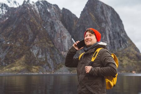 Portrait traveler man talking on mobile phone. Tourist in a yellow backpack standing on a background of a mountain and a lake. Traveler walks, smiling man calling friends. Telephone conversation.の写真素材
