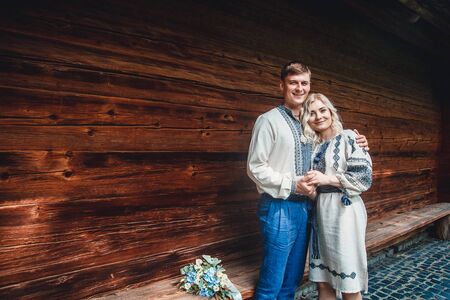 Amazing wedding couple in an embroidered shirt with a bunch of flowers on the background of a wooden houseの写真素材
