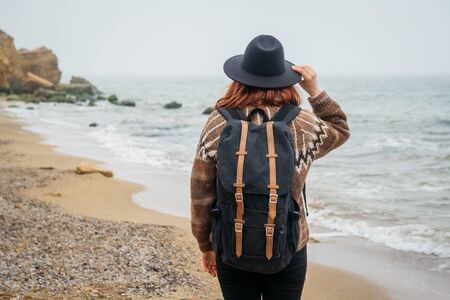 Portrait of a young red-haired woman in a hat and a scarf with a backpack against the background of the rocks against the beautiful sea, on the coast, on the horizon. Tourism, rest, lifestyle. Shoot from the backの写真素材