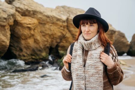 Portrait of a young red-haired woman in a hat and a scarf with a backpack against the background of the rocks against the beautiful sea, on the coast, on the horizon. Tourism, rest, lifestyle.の写真素材