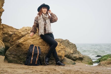 Portrait of a young red-haired woman in a hat and a scarf with a backpack against the background of the rocks against the beautiful sea, on the coast, on the horizon. Tourism, rest, lifestyle.の写真素材