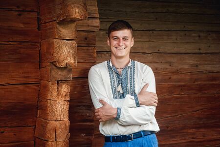 Beautiful young guy in an embroidered shirt on the background of a wooden houseの写真素材