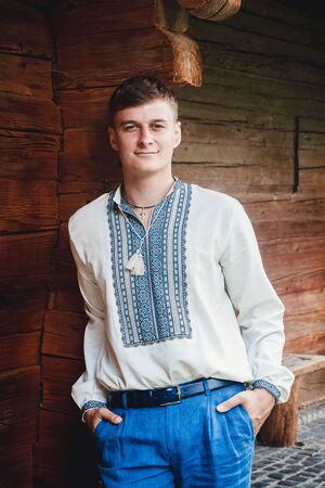 Beautiful young guy in an embroidered shirt on the background of a wooden houseの写真素材