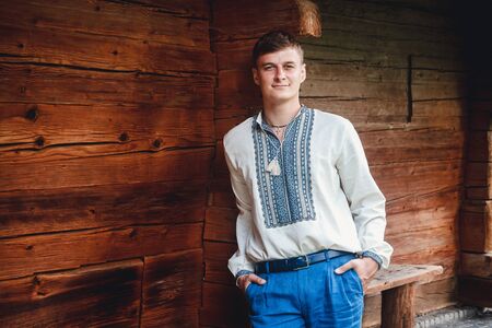 Beautiful young guy in an embroidered shirt on the background of a wooden houseの写真素材