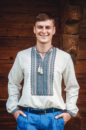 Beautiful young guy in an embroidered shirt on the background of a wooden houseの写真素材