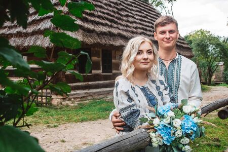 Amazing wedding couple in an embroidered shirt with a bunch of flowers on the background of a wooden houseの写真素材
