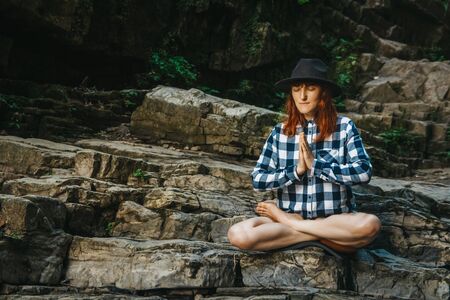 Beautiful girl with red hair in a hat and shirt meditating on rocks in a lotus position. Space for your text message or promotional contentの写真素材