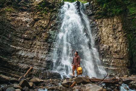 Traveler man with a yellow backpack standing on the background of a waterfall. Travel lifestyle concept.の写真素材