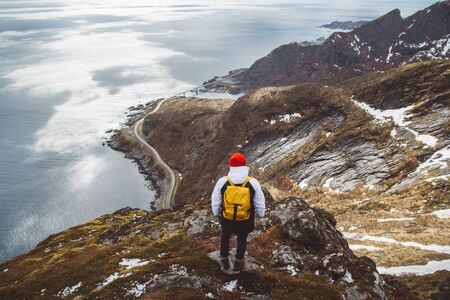 Traveler man with a yellow backpack wearing a red hat standing on the background of mountains and sea. Travel lifestyle concept. Shoot from the back.の写真素材