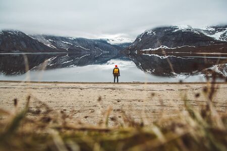 Traveler man with a yellow backpack wearing a red hat standing on the background of mountains and lake enjoying landscape in Lofoten, Norway. Travel lifestyle concept. Shoot from the backの写真素材