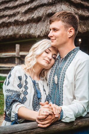Amazing wedding couple in an embroidereds shirt with a bunch of flowers on the background of a wooden houseの写真素材