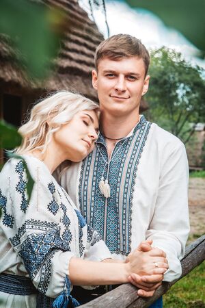 Amazing wedding couple in an embroidereds shirt with a bunch of flowers on the background of a wooden houseの写真素材