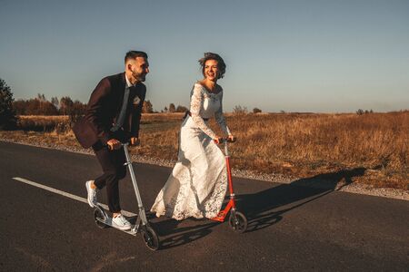 Smiling wedding couple riding a on scooters along the road outside the city at sunset.の写真素材