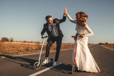 Smiling wedding couple riding a on scooters along the road outside the city at sunset.の写真素材