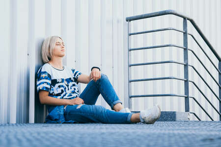 Beautiful girl with short white hair dressed in jeans in urban style sits on the background of a white wall. Place for text.の写真素材