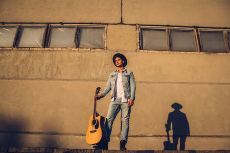 Young guy musician in a hat with a guitar against a wall of concreteの写真素材